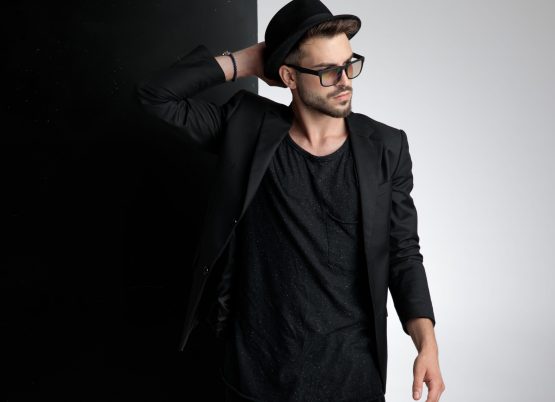 attractive young casual man wearing black hat, black coat and sunglasses, holding hand to head and looking to side, standing on black and white background in studio